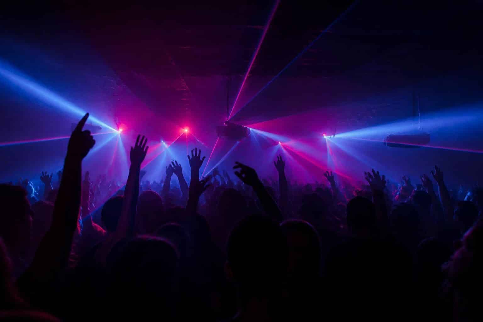 Clubbers with hands raised under pink and blue lasers in a dark house music club